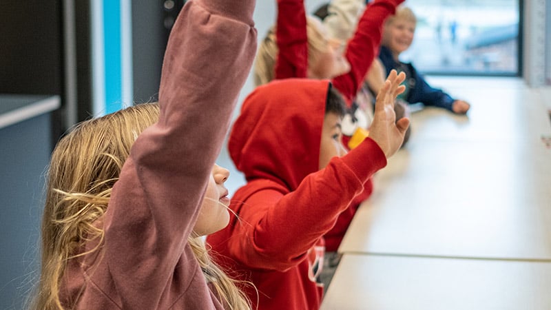 Elever sitter längs ett bord i klassrum och räcker upp händerna